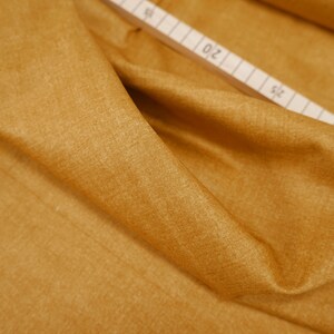 Peut inclure: Gros plan sur un tissu jaune moutarde avec une texture subtile. Le tissu est plié et drapé, révélant sa douceur et sa fluidité.