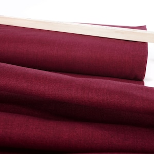 Könnte beinhalten: Eine Rolle tiefroter Stoff mit einer dezenten Textur. Der Stoff ist gefaltet und über einen Holzstab gelegt.