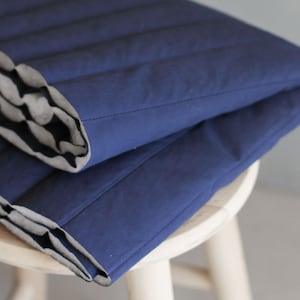 Könnte beinhalten: Eine dunkelblaue gesteppte Wolldecke, die auf einem Holzhocker gefaltet ist. Die Decke hat einen weißen und schwarzen Streifenbesatz.