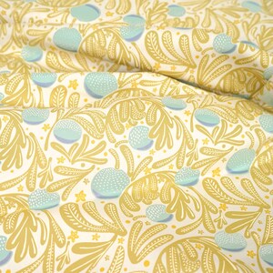 Könnte beinhalten: Ein weißer Stoff mit einem sich wiederholenden Muster aus goldenen Blättern und blauen und weißen Kreisen. Der Stoff ist gefaltet und über ein Holzlineal gelegt.