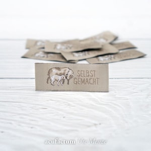 May include: A beige fabric label with a brown sheep and lamb illustration and the text "SELBST GEMACHT".