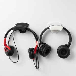 May include: Two pairs of headphones hanging on a white wall. One pair is black with red accents, the other is all black. The headphones are mounted on black and white wall hooks.
