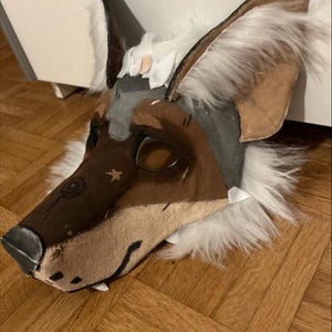 May include: A handmade, stylized wolf mask with brown, gray, and white fur accents. The mask features large, pointed ears, a detailed snout, and eye openings. The mask is designed for costume or cosplay use.
