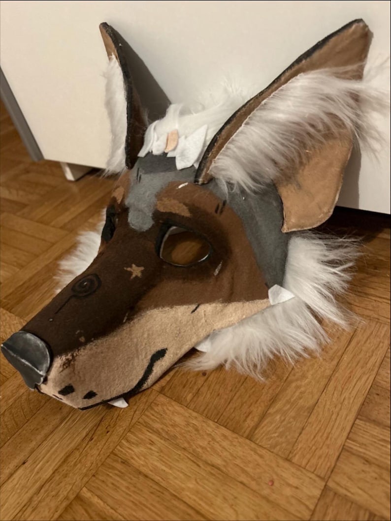 May include: A handmade, stylized wolf mask with brown, gray, and white fur accents. The mask features large, pointed ears, a detailed snout, and eye openings. The mask is designed for costume or cosplay use.