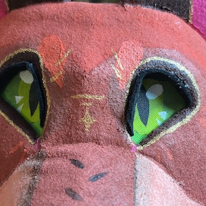 May include: Close-up of a handmade red and pink furry mask with green eyes. The mask has gold accents and a brown and pink ear design. The eyes are a bright green with black detailing.