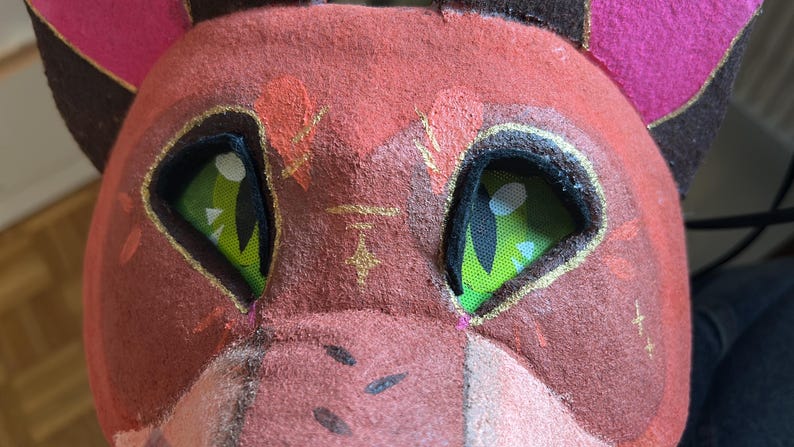 May include: Close-up of a handmade red and pink furry mask with green eyes. The mask has gold accents and a brown and pink ear design. The eyes are a bright green with black detailing.