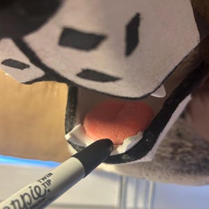 May include: A close-up of a black and white Sharpie Twin Tip marker pointing towards the inside of a plush animal head. The animal head has a pink tongue and white teeth.