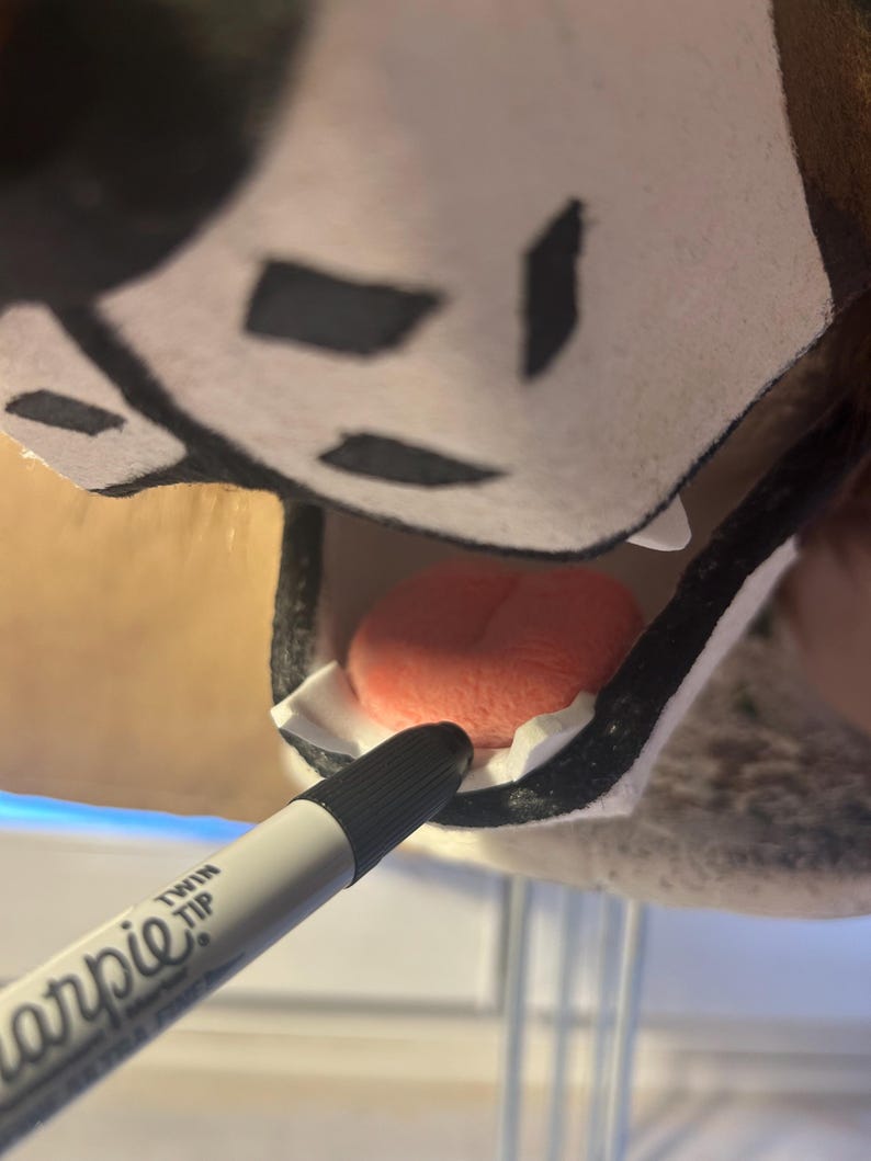 May include: A close-up of a black and white Sharpie Twin Tip marker pointing towards the inside of a plush animal head. The animal head has a pink tongue and white teeth.