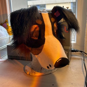 May include: A handmade dog head, featuring a white muzzle, black and brown spots, and black fur around the ears. The dog head is resting on a light gray base, with a black nose and small black dots.