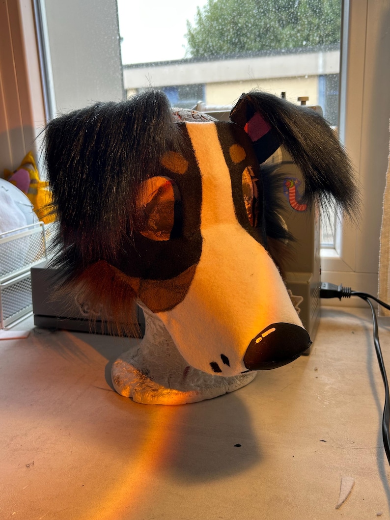 May include: A handmade dog head, featuring a white muzzle, black and brown spots, and black fur around the ears. The dog head is resting on a light gray base, with a black nose and small black dots.