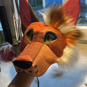 May include: A handcrafted fox mask with orange, brown, and white fur details. The mask features green mesh eyes, a pointed snout, and large, upright ears. The mask is held by a person's hand.