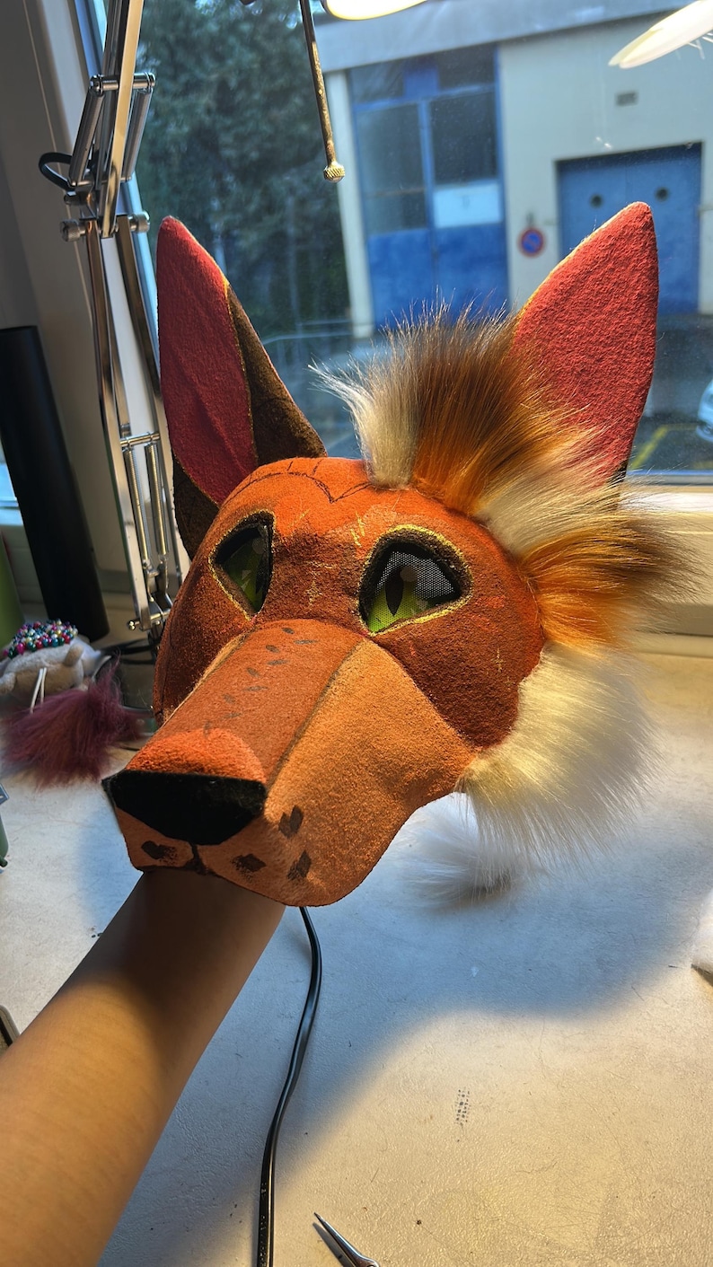 May include: A handcrafted fox mask with orange, brown, and white fur details. The mask features green mesh eyes, a pointed snout, and large, upright ears. The mask is held by a person's hand.