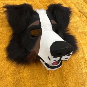 May include: A dog-themed mask with black, white, and brown fur. The mask features a black nose, white muzzle, and brown accents around the eyes. The ears are black and fluffy. The mask is designed to be worn.