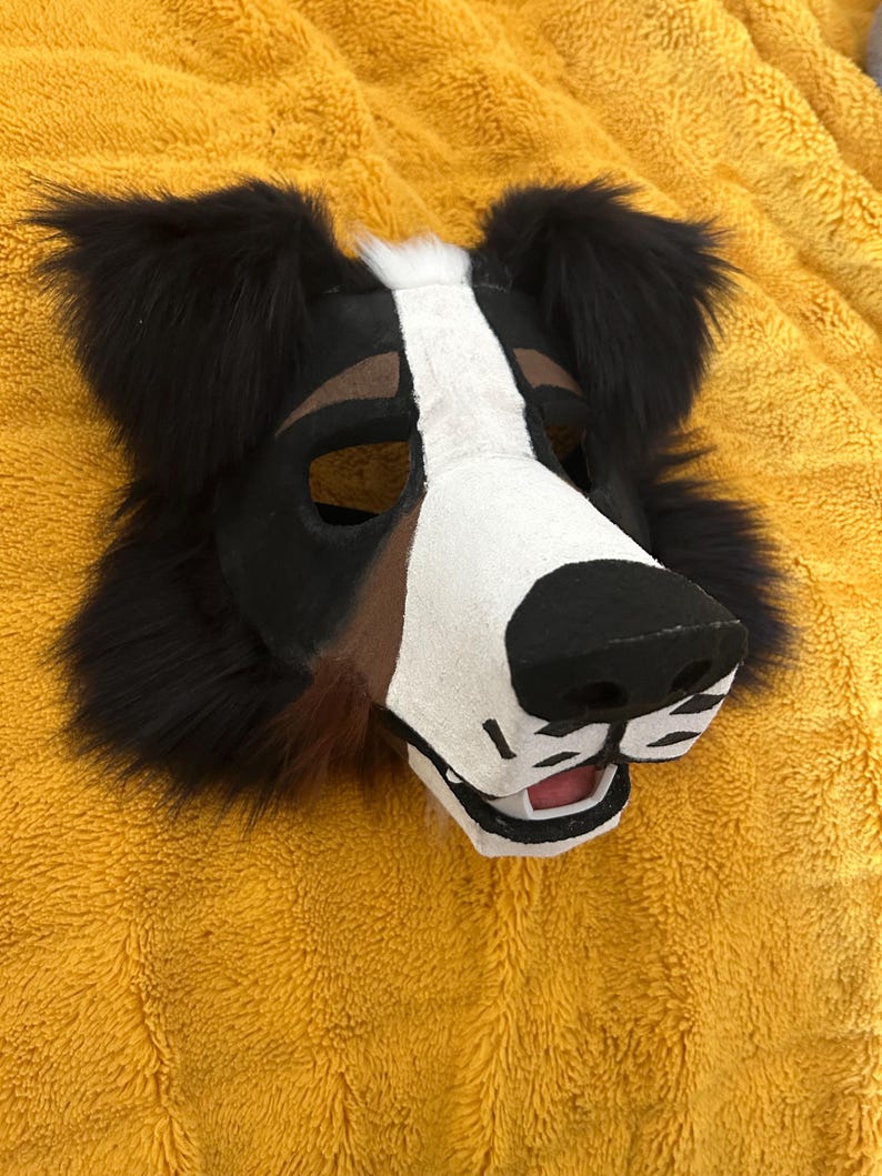 May include: A dog-themed mask with black, white, and brown fur. The mask features a black nose, white muzzle, and brown accents around the eyes. The ears are black and fluffy. The mask is designed to be worn.