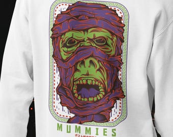 May include: A white hooded sweatshirt featuring a graphic on the back of a green and purple mummy head wrapped in bandages. The design is enclosed in a rectangular border with a Greek key pattern and includes the text MUMMIES in green letters.