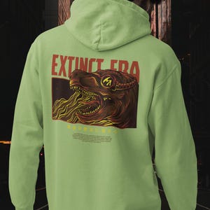 May include: A light green hooded sweatshirt featuring a graphic on the back. The design displays a dinosaur head in shades of brown and yellow with the text "EXTINCT ERA" in red letters above it. The hoodie has long sleeves and a drawstring hood.