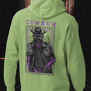 May include: A light green hooded sweatshirt featuring a graphic on the back. The design shows a bull wearing a cowboy hat, a dark vest, and a sheriff badge, framed by a rectangular border with the text "WARRIOR COWBOY" in purple lettering.