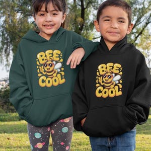 Könnte beinhalten: Zwei Kinder in passenden Kapuzenpullovern mit einem "Bee Cool"-Motiv. Ein Kapuzenpullover ist dunkelgrün, der andere schwarz. Das Mädchen trägt gemusterte Leggings, und der Junge trägt blaue Jeans. Sie stehen auf Gras vor Bäumen.