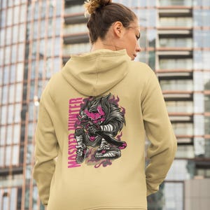 May include: A beige hooded sweatshirt featuring a graphic on the back of a stylized character with a pink face and dark hair holding a sword. The text "MASTER HUNTER" is printed vertically in pink next to the character, surrounded by purple smoke.