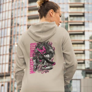 May include: A beige hooded sweatshirt featuring a graphic print on the back. The design depicts a stylized character with a pink face and dark hair holding a sword, accompanied by the vertical text "MASHER HUNTER" in bold pink capital letters.
