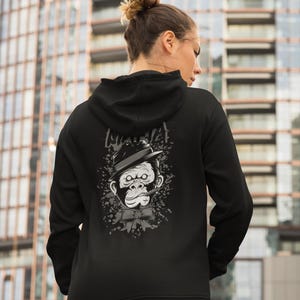 May include: A black hooded sweatshirt featuring a graphic print on the back. The design displays a grayscale illustration of a gorilla wearing a fedora hat and a bow tie, with a cigar in its mouth, surrounded by a splattered ink effect.