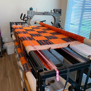 May include: A longarm quilting machine with a large orange and black quilt on the frame. The machine is a Gammill and has a pink measuring tape attached to the frame.