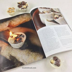 Pu&ograve; includere: Un primo piano di una pagina di rivista con una ricetta per fare candele. La pagina mostra una foto di diverse piccole candele bianche con noci e gocce di cioccolato incorporate nella cera. La rivista si chiama "GreenCraft" e il numero di pagina &egrave; 54. La rivista &egrave; aperta a una pagina con testo su come fare le candele.