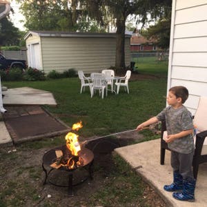 Pu&ograve; includere: Un bambino in una camicia mimetica e stivali di gomma blu arrostisce un marshmallow su un fal&ograve; in un cortile.