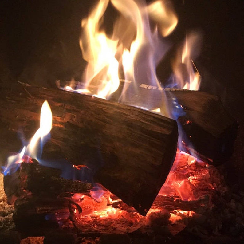 Pu&ograve; includere: Primo piano di un camino con ceppi ardenti. Le fiamme sono arancioni e gialle, con alcune fiamme blu visibili. I ceppi sono carbonizzati e hanno una consistenza ruvida.