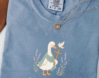 May include: Blue jean-colored t-shirt with a graphic of a goose holding a cross and a dove. The shirt has the words "Faith Mode On" printed on it. A gold necklace with a pendant is draped across the shirt.