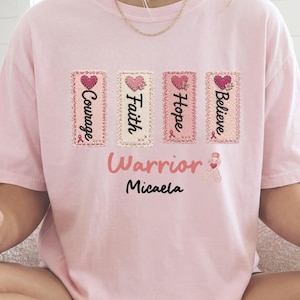 May include: A light pink t-shirt featuring four vertical rectangular patches with the words Courage, Faith, Hope, and Believe, each topped with a pink heart. Below these, the word Warrior is printed in pink, followed by the name Micaela in black.