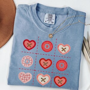 May include: Blue jean colored t-shirt with a tic-tac-toe design featuring red hearts and circles with brown cross and circle accents. A gold necklace with a pendant is draped across the shirt. The shirt is labeled "Comfort Colors".