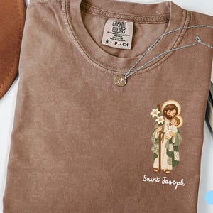 May include: A brown short-sleeved t-shirt with a crew neckline and a Comfort Colors brand label. The shirt features a graphic of Saint Joseph holding a child and a lily, with the text Saint Joseph printed below in white script lettering.