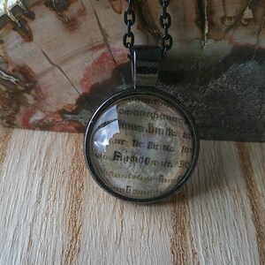 May include: A silver necklace with a round glass pendant featuring a black and white image of a page from an old book. The text on the page is in a gothic font.