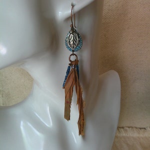 May include: A pair of dangle earrings with a blue and silver leaf charm and a brown fabric tassel.