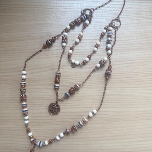 May include: A multi-strand necklace with copper-colored chains and beads. The necklace features white, brown, and blue beads, along with a circular pendant with a cross design. The necklace is displayed on a light brown wooden surface.