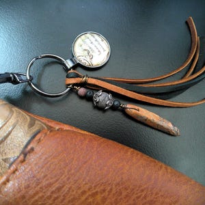 May include: A handmade keychain with a silver ring, a round charm with script, and leather tassels in black and brown. The keychain also features a decorative stone and a wooden accent, attached to a brown leather wallet.
