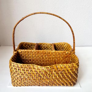 May include: A woven rattan basket with a handle. The basket is divided into three compartments and has a rectangular shape. The basket is a warm brown color.