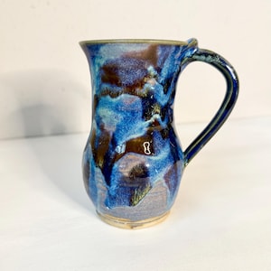 May include: Handmade ceramic mug with a unique, organic shape. The mug features a dark blue glaze with brown and lighter blue accents, creating a marbled effect. The handle is a matching dark blue. The mug is suitable for hot or cold beverages.