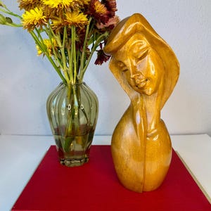 May include: A wooden sculpture of a woman's head and shoulders with closed eyes. The sculpture is light brown and sits on a red book. A vase of yellow and maroon flowers is to the left.