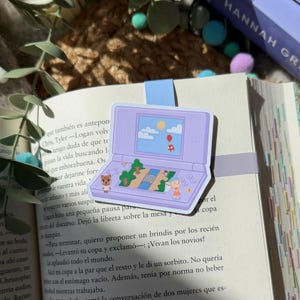 Könnte beinhalten: Ein lila Handheld-Videospiel-Aufkleber mit einer Landschaftsszene, Zeichentrickfiguren, Bäumen und einem Ballon. Der Aufkleber befindet sich auf einem offenen Buch mit Text.