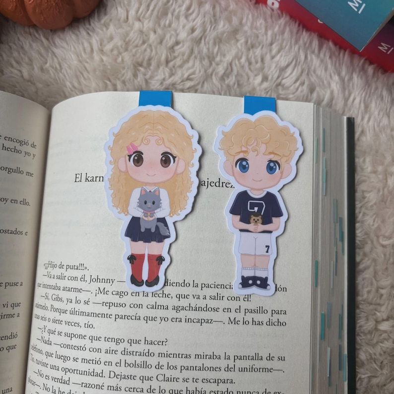 May include: Two anime-style character bookmarks. One shows a girl with blonde curly hair holding a grey cat, the other a boy with blonde hair wearing a dark blue shirt with the number 7. Both are placed in a book, enhancing the reading experience.