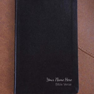 以下が含まれることがあります： 「Your Name Here」と「Bible Verse」の白文字が入った黒い聖書。赤と青のリボンのしおりが付いています。表紙は質感があり、背景は茶色です。