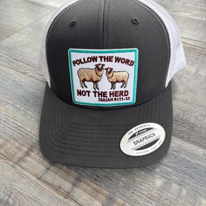 Puede incluir: Gorra de camionero gris y blanca con un parche rectangular con dos ovejas y el texto "FOLLOW THE WORD NOT THE HERD ISAIAH 8:11-13". La gorra tiene una visera curva y un cierre snapback.