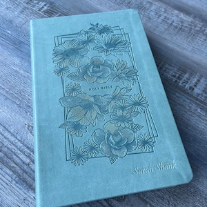 Puede incluir: Una Biblia Sagrada de color azul claro con ilustraciones florales y el texto "Holy Bible" y "Sarah Shank". La portada presenta un marco rectangular con diseños detallados de flores y hojas.