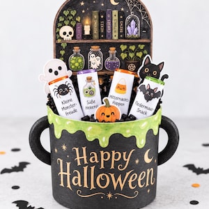 May include: A black cauldron-shaped container with green slime drip detail and the text Happy Halloween. It holds four candy bars labeled Kleine Monsterfreude, Süße Hexerei, Mitternachts-Happen, and Fledermaus-Snack, plus a pumpkin, ghost, and cat cutout.