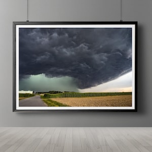 May include: A dark, stormy sky hangs low over a field of green crops. A paved road leads through the field, with a small building visible in the distance.
