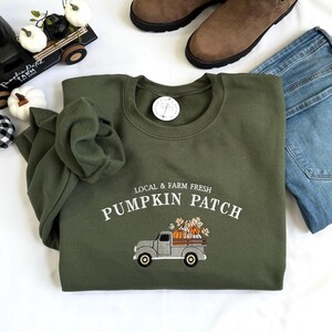 May include: An olive green crewneck sweatshirt featuring white embroidered text that reads LOCAL & FARM FRESH PUMPKIN PATCH above a graphic of a gray pickup truck carrying orange pumpkins and autumn leaves on the back.