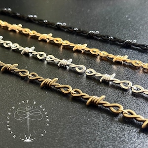 May include: Close-up of four barbed wire necklaces in different metallic finishes: black, gold, silver, and bronze. The necklaces are arranged diagonally against a dark background. The image includes the text "Madison Art & Design Co."