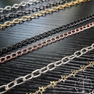 May include: Close-up of various metal chain styles in silver, black, gold, and rose gold. The chains are arranged diagonally, showcasing different link designs, including oval and star-shaped links. The background is a dark wood surface.